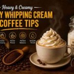 Creamy coffee topped with whipped cream beside heavy whipping cream bottle and coffee beans on dark rustic background